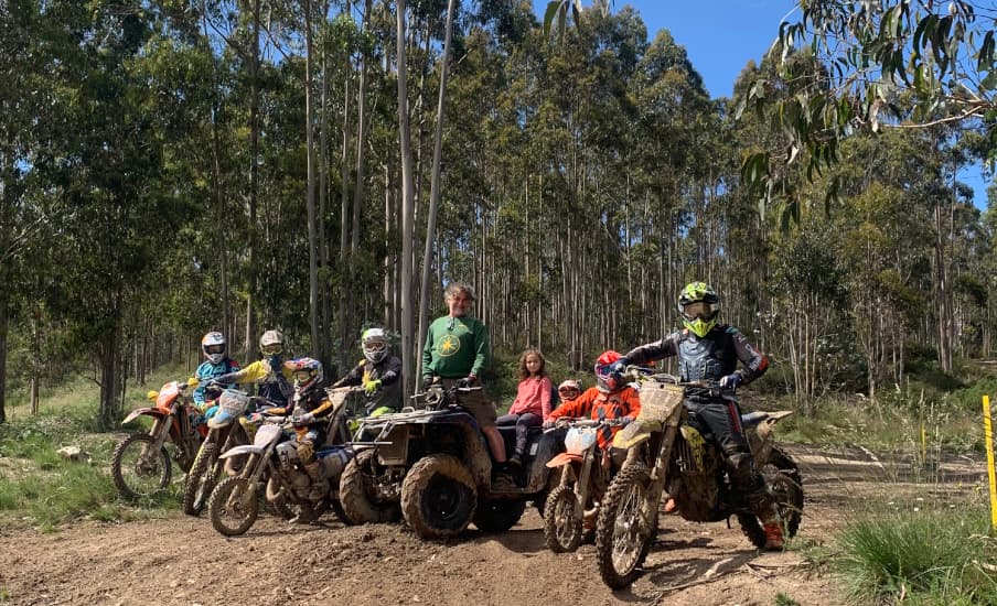 Grupo de niños y adultos posando con sus motos de cross y un quad en una pista de tierra.